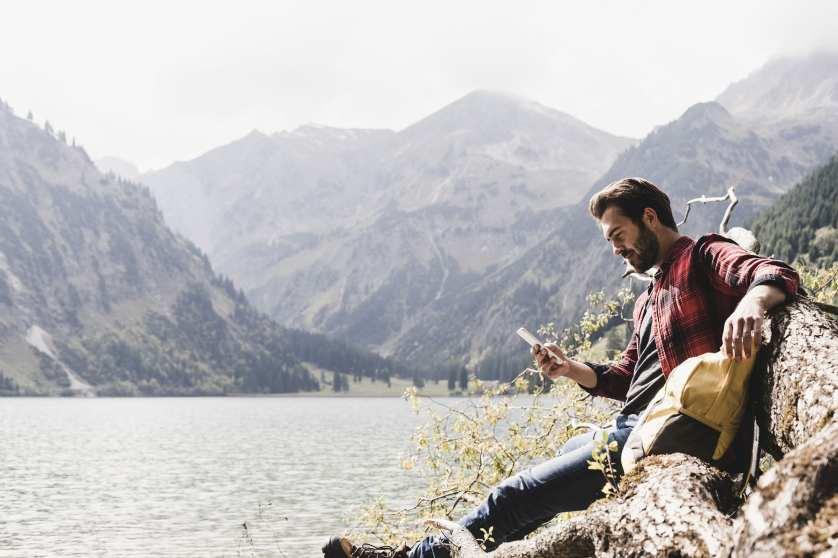 Austria, Tyrol, Alps, tourist hiker relaxing on tree trunk at mountain lake checking cell phone 