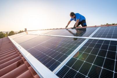 Kneeling professional fixing solar panels from the top of a house roof, side view of the roof with sun reflection
